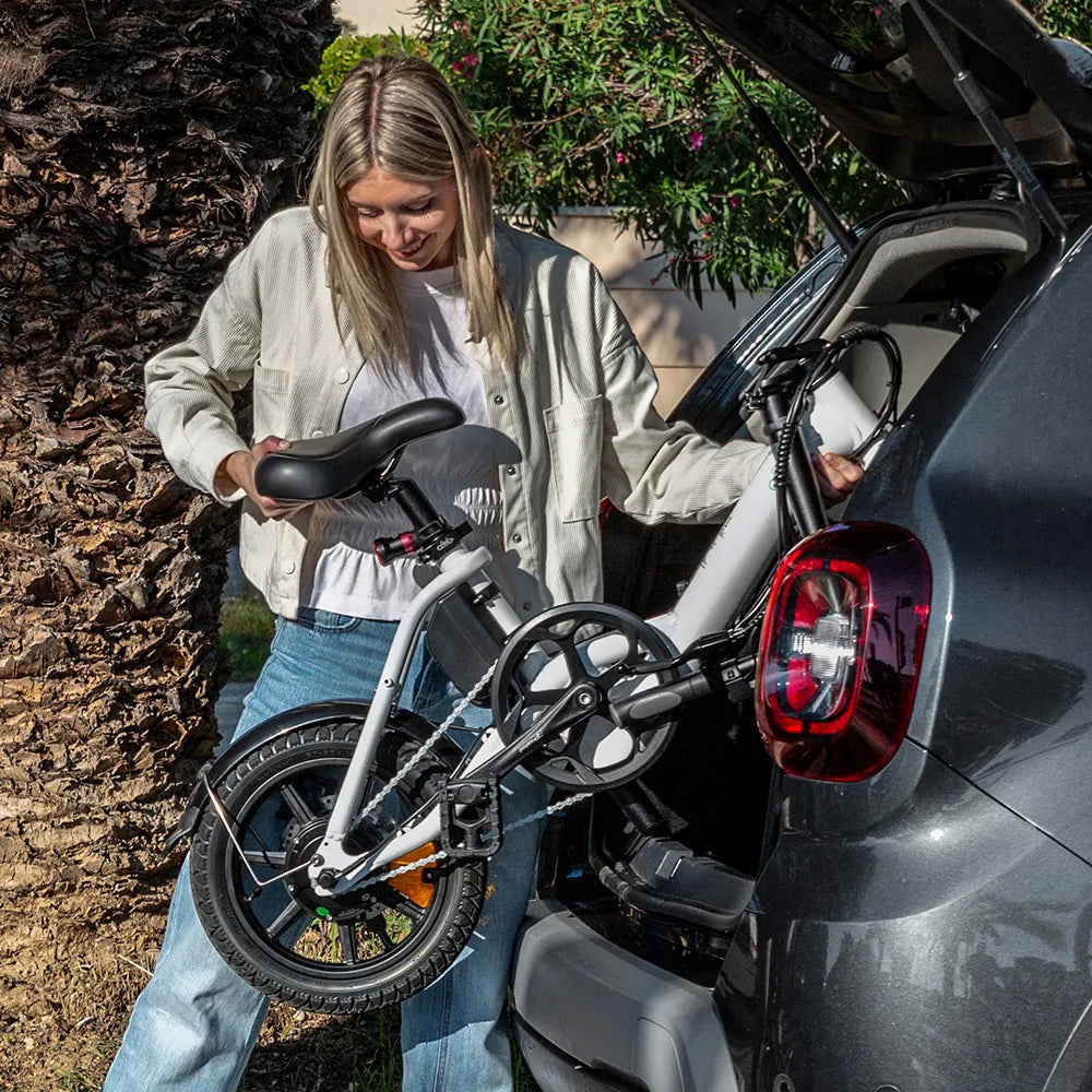 Nainen hymyilee nostaessaan SÄHKÖPYÖRÄT.NET Fiido D3 Pro Mini taitettava sähköpyörä autonsa takakontista, taustalla rehevää vehreyttä ja puita.