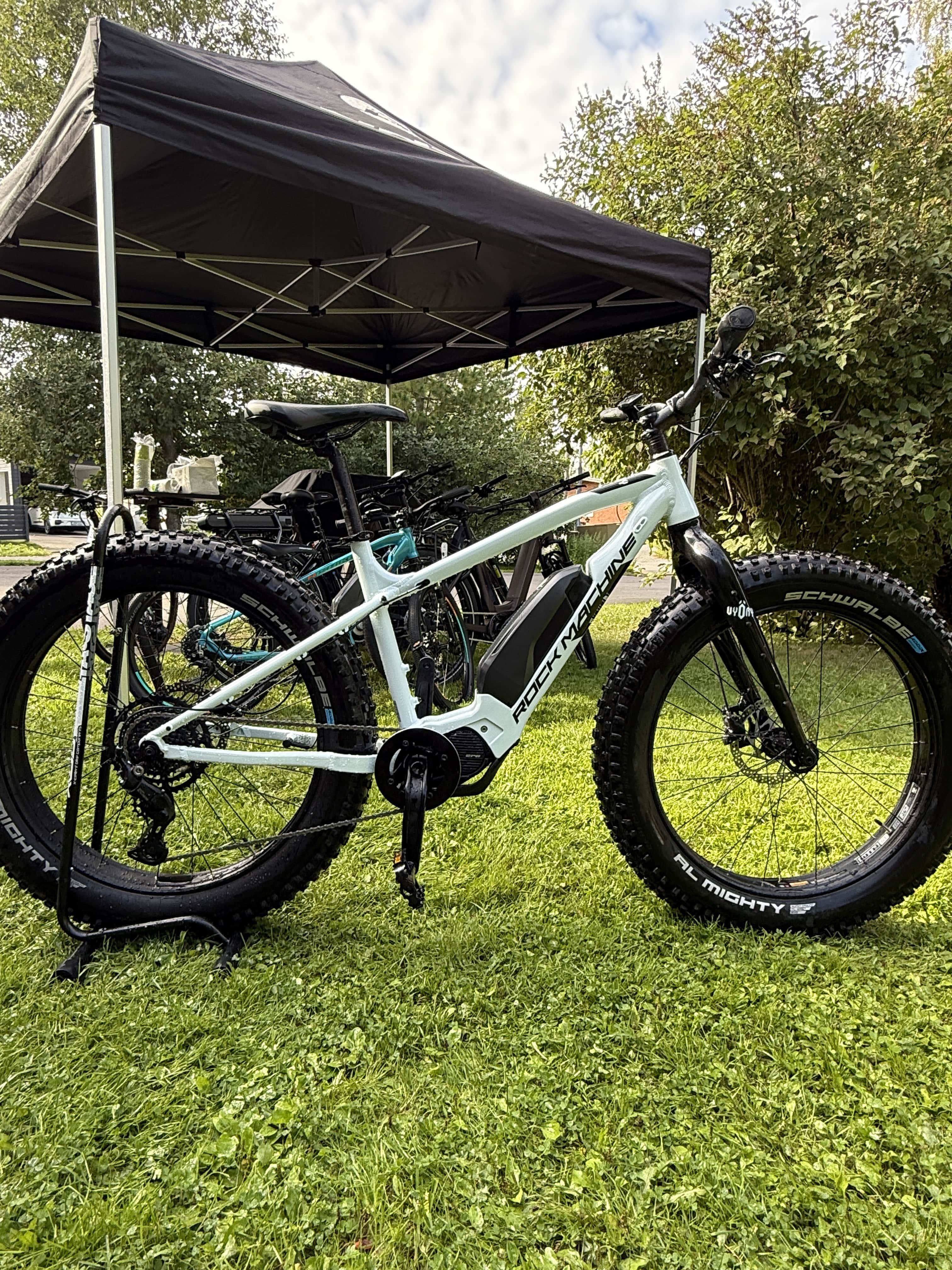 Valkoinen Rock Machine E50R sähköpyörä (ajettu 120-160 km) by SÄHKÖPYÖRÄT.NET, jossa on Maxxis Minion 26x4.80 renkaat, on esillä ruohikolla mustan katoksen alla, ja taustalla on puita ja toinen pyörä.