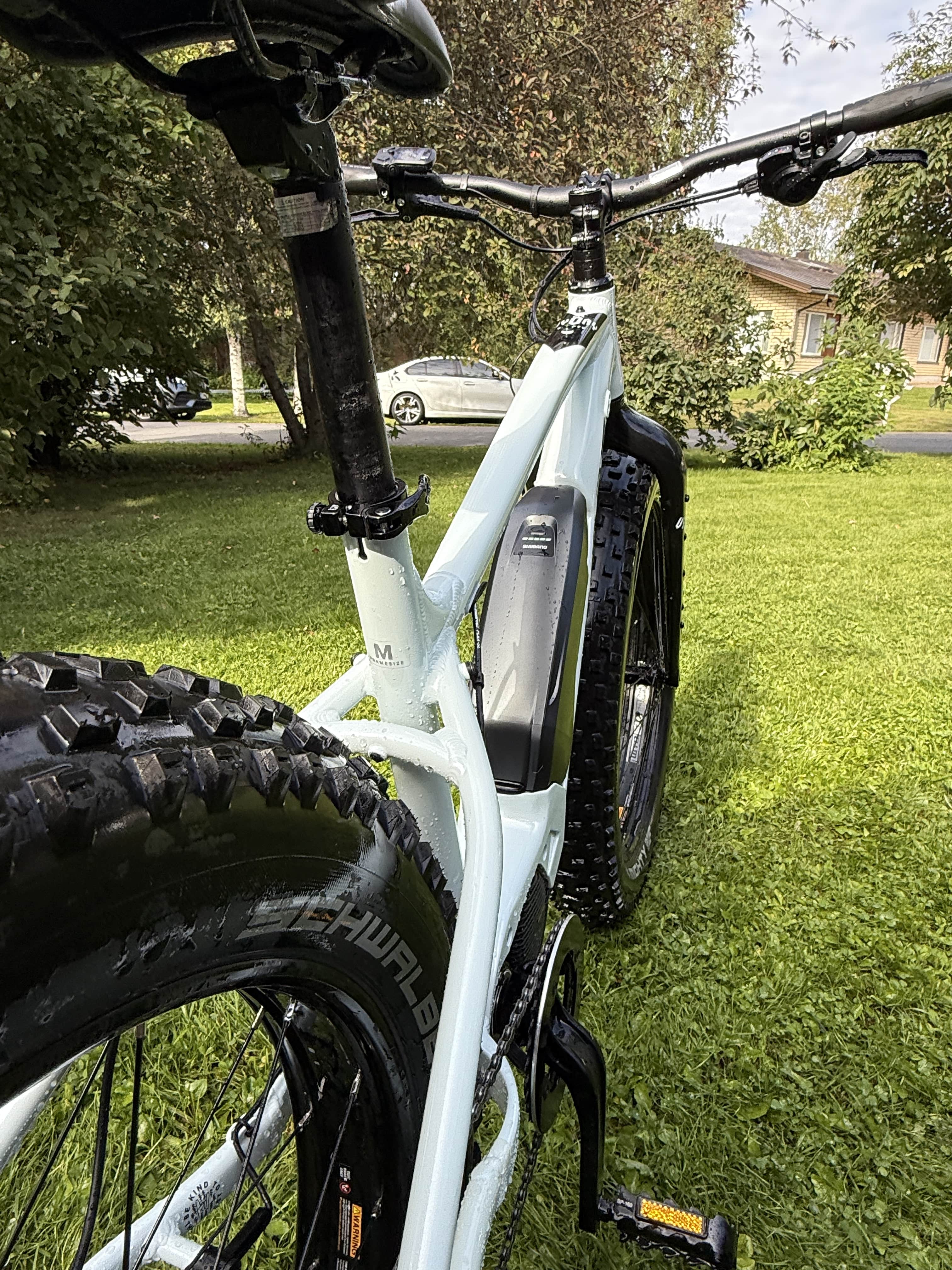 Lähikuva valkoisesta Rock Machine E50R sähköpyörästä (SÄHKÖPYÖRÄT.NET), jossa on Maxxis Minion 26x4.80 takarenkaat ja näkyvä akku, pysäköity ruohikolle; taustalla puita ja talo pehmeästi sumeina. Ajettu 120-160 km.