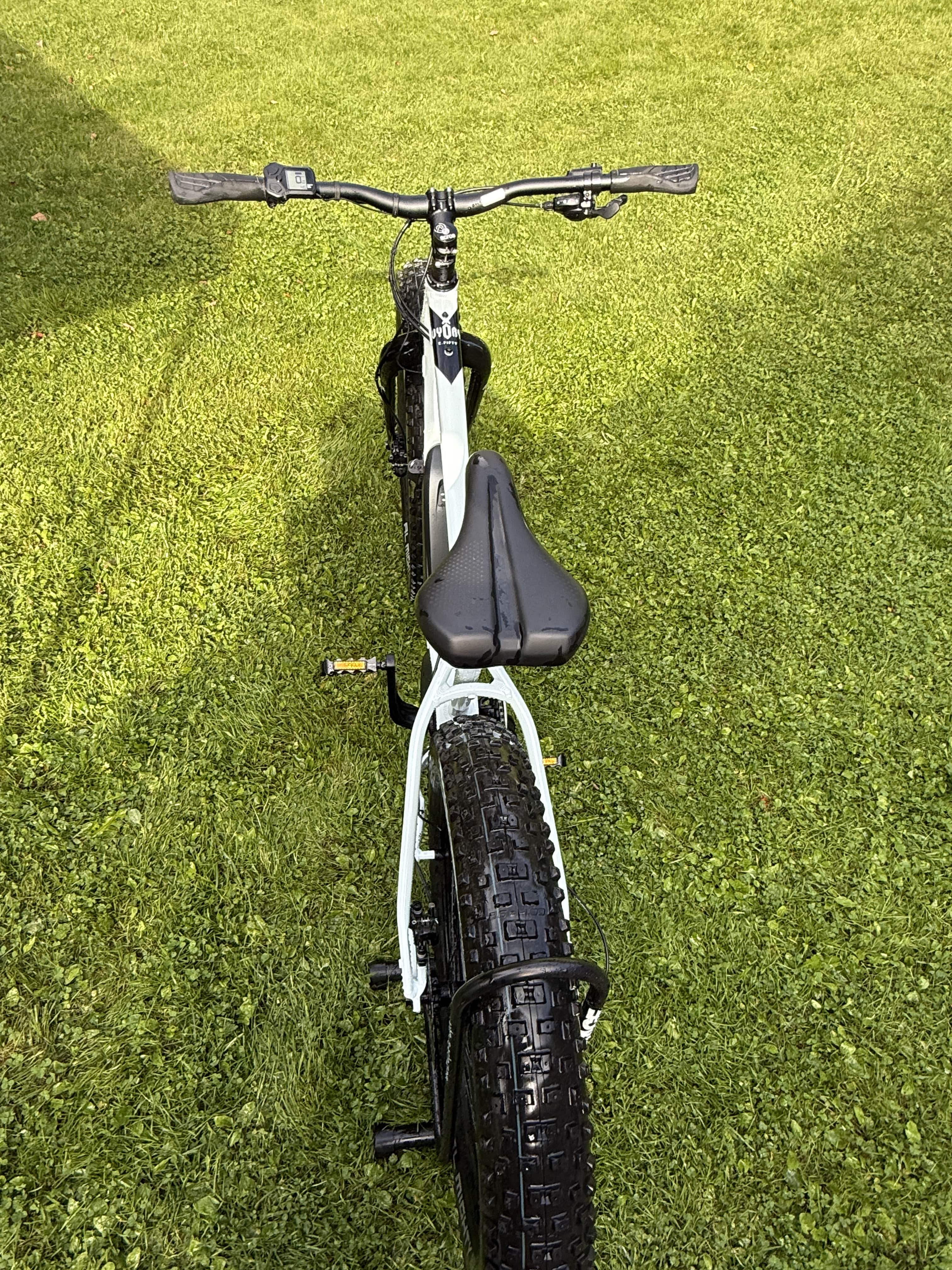 SÄHKÖPYÖRÄT.NET Rock Machine E50R esittelypyörä, ajettu 120-160 km, mustalla istuimella ja ohjaustangolla, on parkkeerattu vihreälle nurmikolle, ja sen leveä Maxxis Minion 26x4.80 -taajuuspyörä ja tyylikäs valkoinen runko näkyvät takaa.