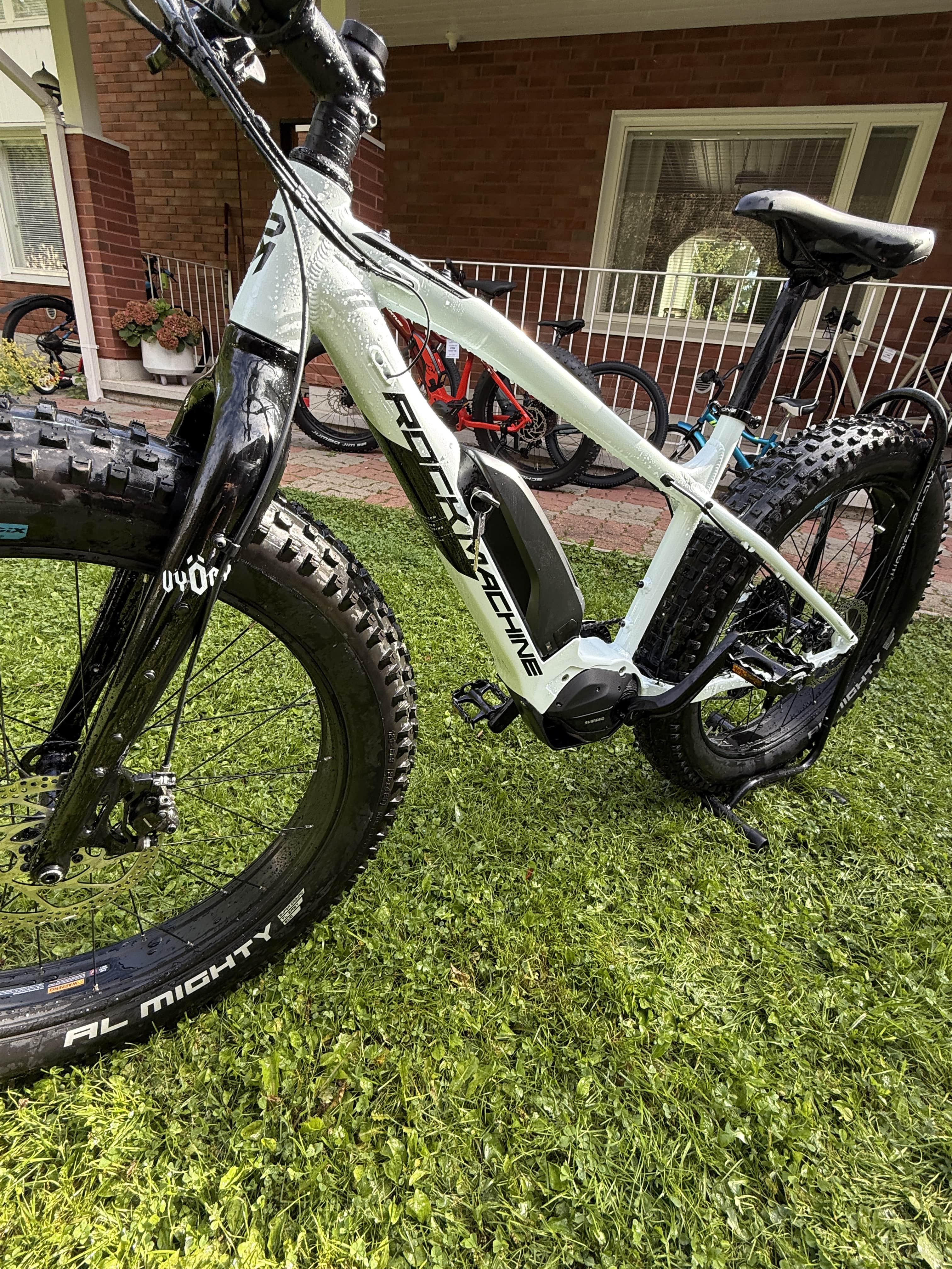 Vaaleansininen Rock Machine E50R esittelypyörä (ajettu 120-160 km), saatavana SÄHKÖPYÖRÄT.NETistä, Maxxis Minion 26x4.80 -renkailla on pysäköity nurmikolle talon lähelle; kuistilla näkyy myös punainen pyörä.