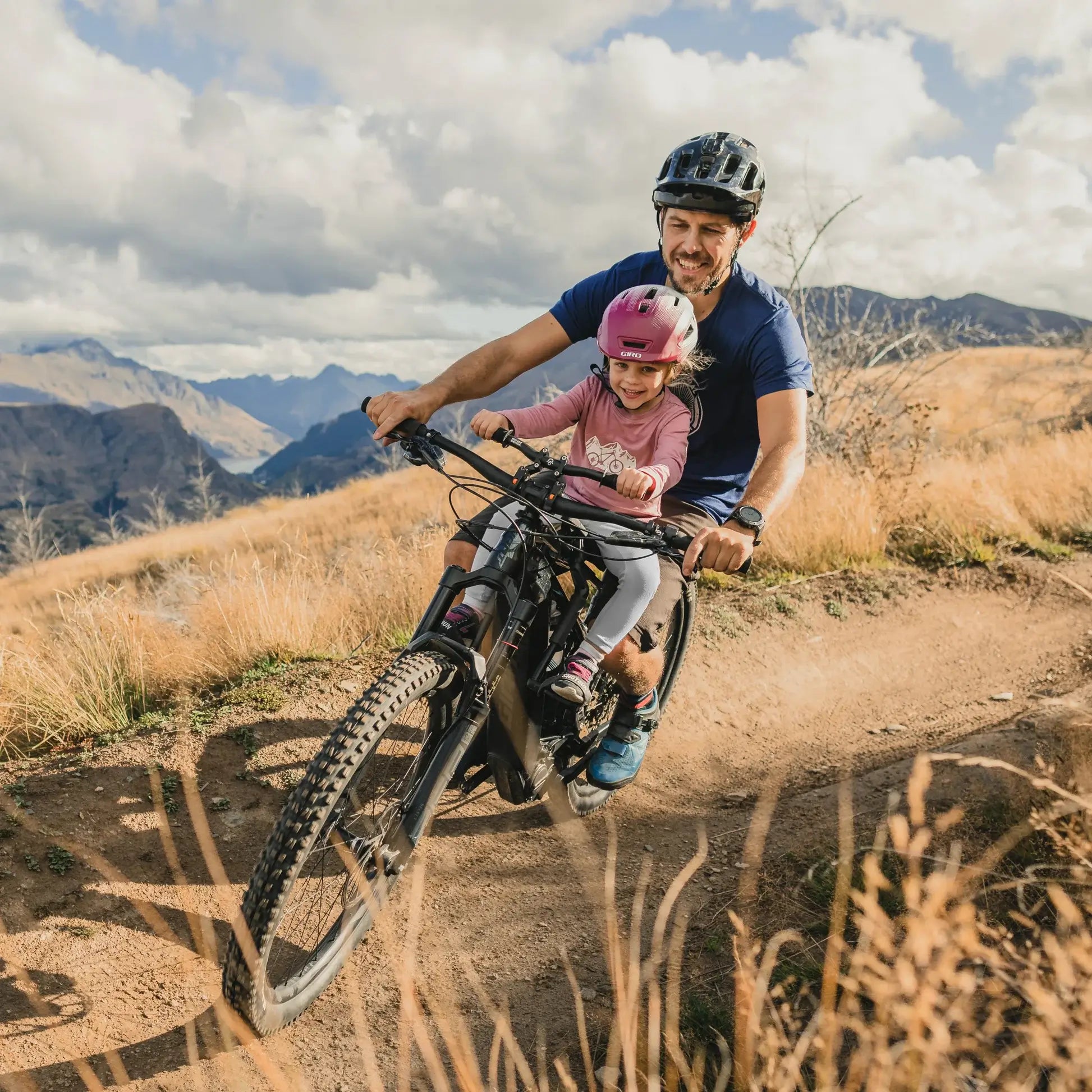 Aikuinen ja pieni lapsi, molemmat kypärä päässä, ajavat maastopyörällä, joka on varustettu SÄHKÖPYÖRÄT.NET SHOTGUN PRO EVO 2IN1 TANKO- JA ISTUINPAKETTI -laitteella, maastopyöräilyreitillä ruohoisilla kukkuloilla, taustalla vuoret ja pilvet.