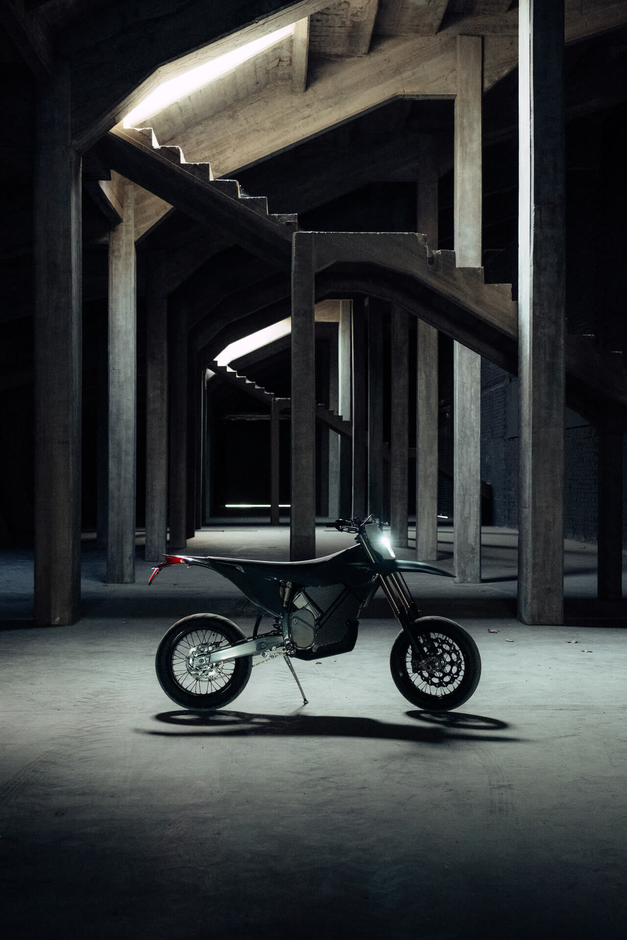 A Stark Varg SM *korkotarjous alkaen 1,99%* sähkömoottoripyörä seisoo ajovalo päällä tyhjän, varjoisan betonirakennelman keskellä tukipilareiden ja ylätasanteiden ympäröimänä.