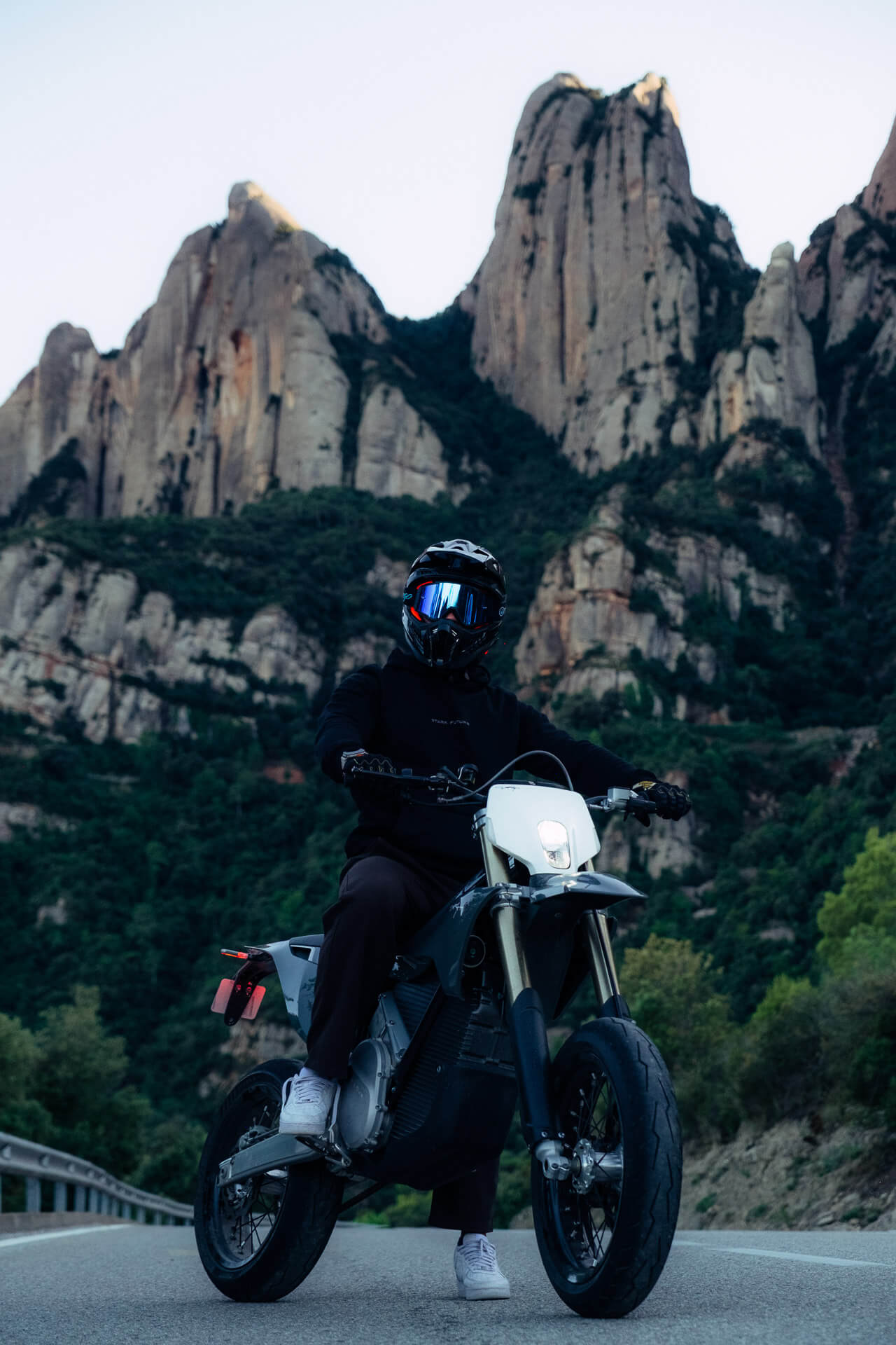 Kypärä ja tummiin vaatteisiin pukeutunut henkilö istuu Stark Varg SM *korkotarjous alkaen 1,99 %* Stark-sähkömoottoripyörän päällä, joka on pysäköity tielle, jonka taustalla on korkeita vuoria ja vihreitä puita.