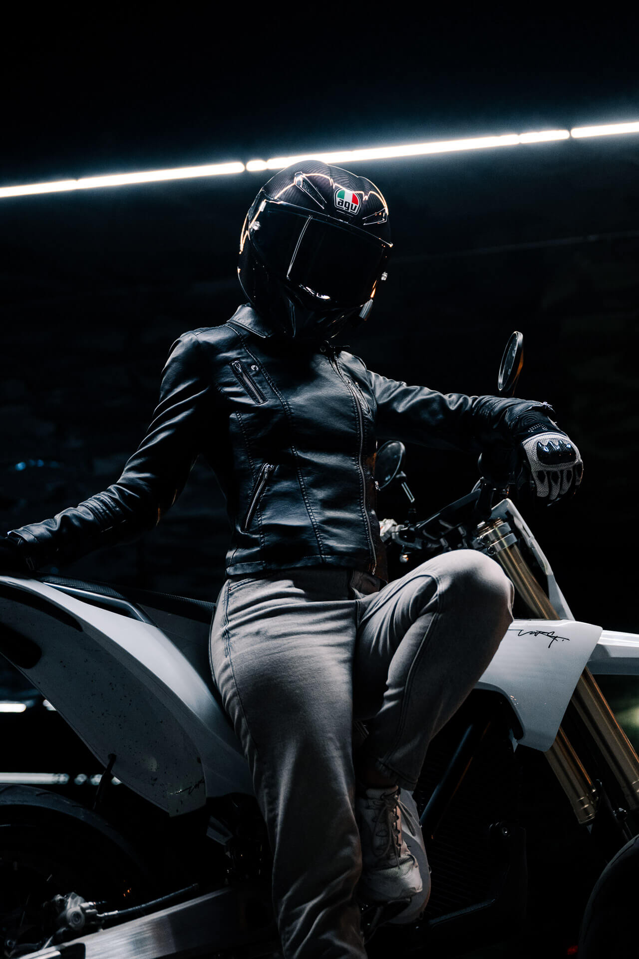 Mustaan kypärään, nahkatakkiin ja harmaisiin housuihin pukeutunut henkilö poseeraa yhdellä jalalla Stark Varg SM *korkotarjous alkaen 1,99 %* sähkömoottoripyörä by Stark dramaattisessa valaistuksessa pimeässä ympäristössä.