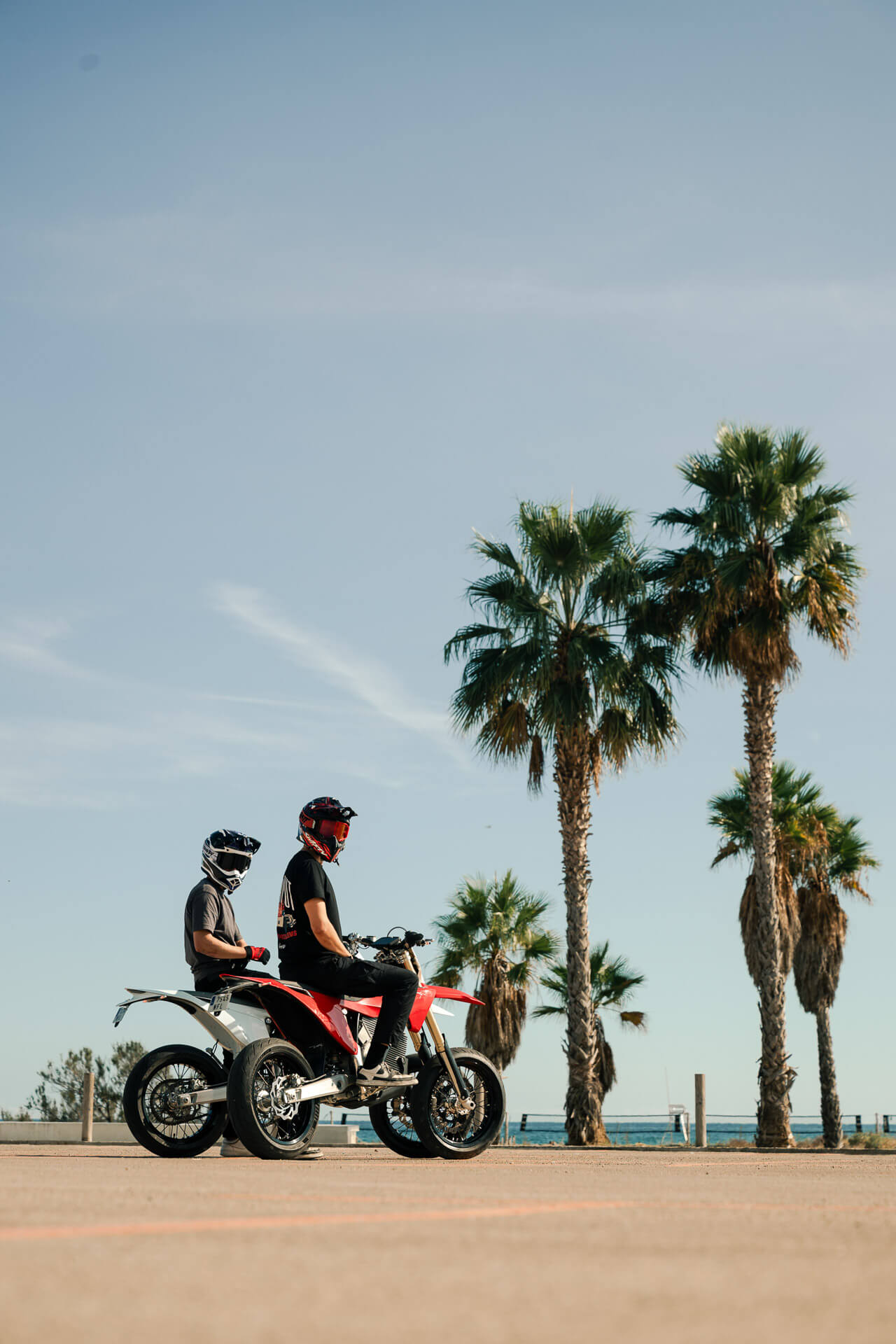 Kaksi kypäräpukuista henkilöä ajaa punamustalla Stark Varg SM supermoto-mönkijällä Starkilla aurinkoisena päivänä, taustalla korkeat palmupuut ja kirkkaan sininen taivas. *Korkotarjous alkaen 1,99 %*.