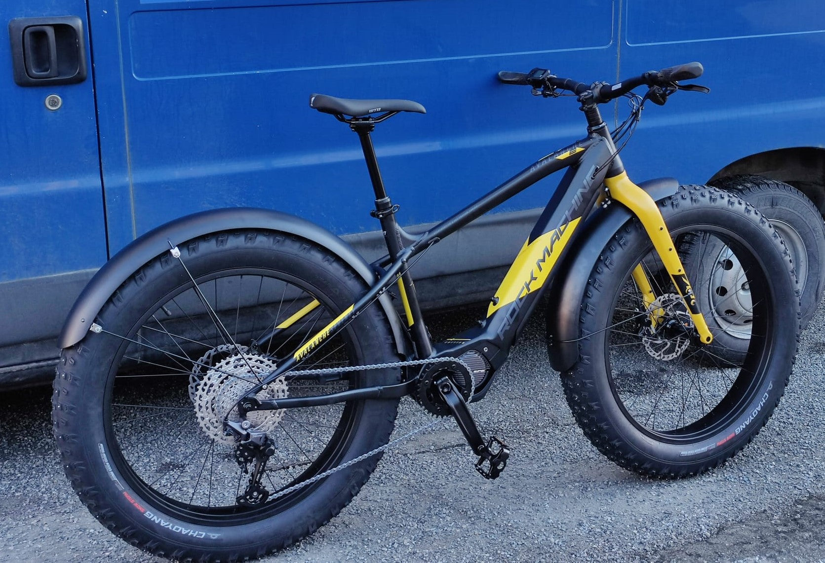 SÄHKÖPYÖRÄT.NETin keltainen ja musta läskirengasmaastopyörä, jossa on FG FATBIKE LOKASUOJASARJA TÄYSPITKÄ 4,8", on pysäköity soralle sinisen pakettiauton eteen. Siinä on erittäin leveät renkaat, täyspitkät lokasuojat ja järeä runko karkeaan maastoon.