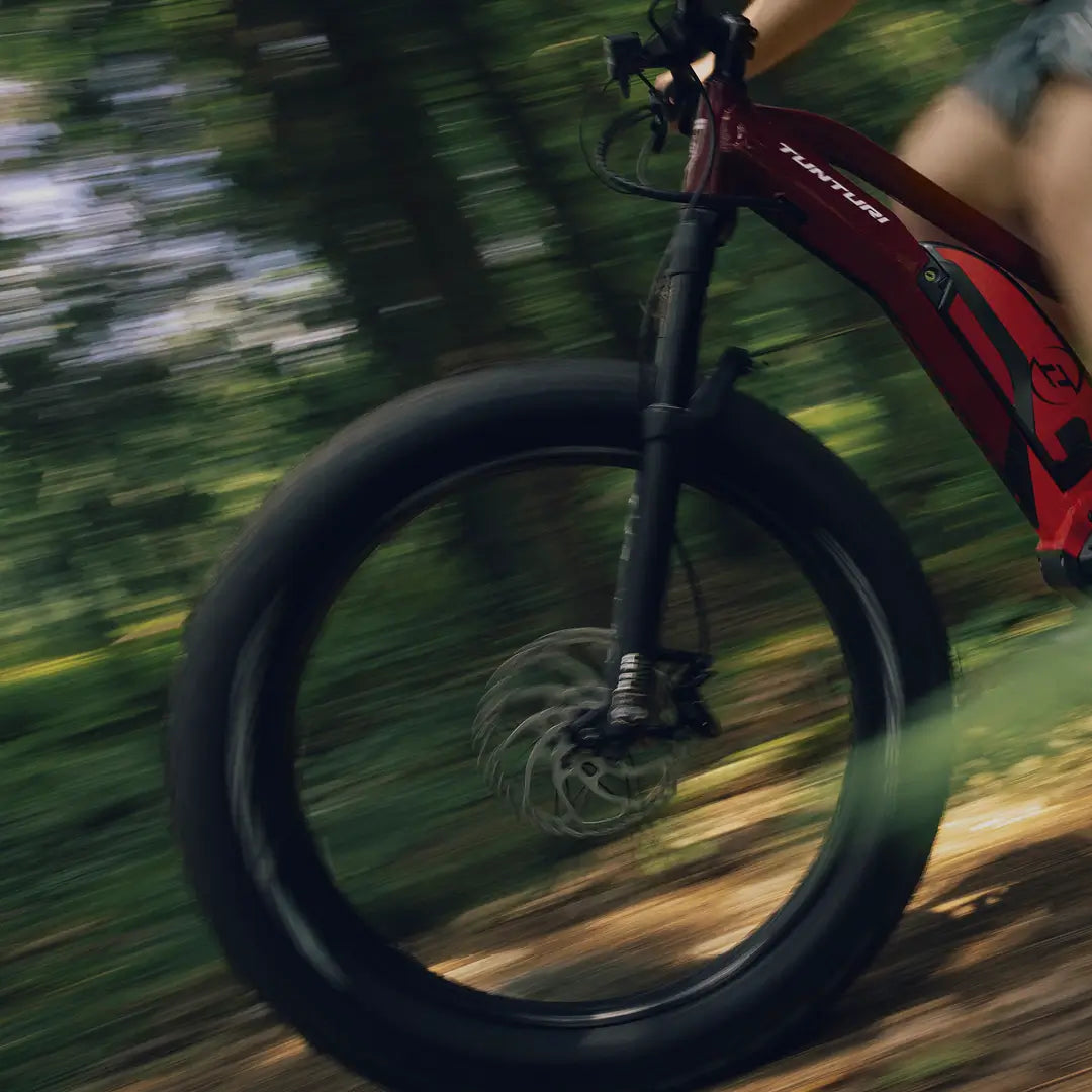 Lähikuva Tunturi EMAX *SUOMEN EDULLISIN* fatbike SÄHKÖPYÖRÄT.NETin paksut renkaat ajavat kilpaa läpi auringonpaisteisen metsän, pyörät ja tausta sumentuvat nopeuden osoittamiseksi.