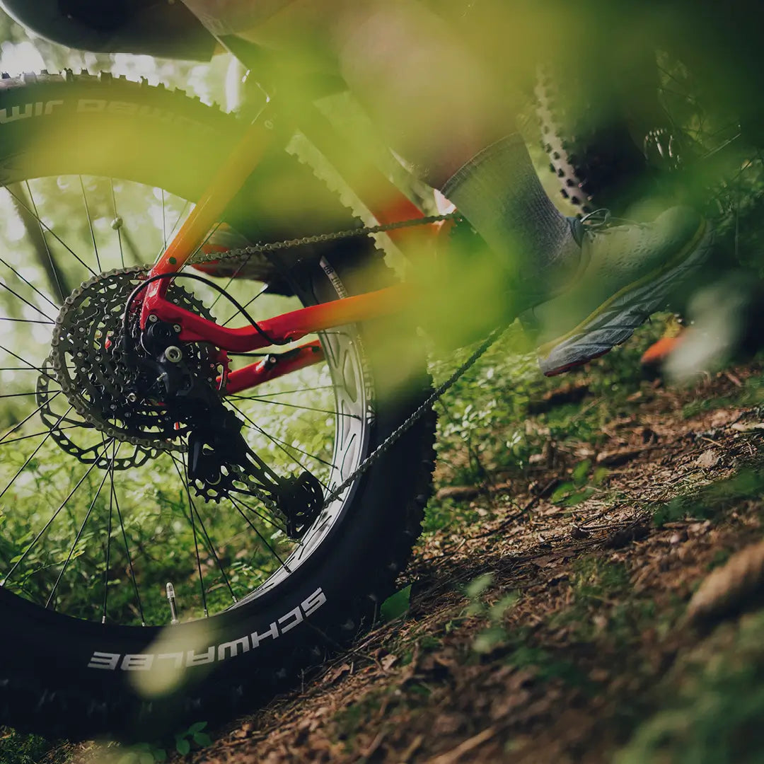 Lähikuva henkilöstä, joka ajaa Tunturi EMAX *SUOMEN EDULLISIN* sähköfatbike -pyörällä metsäpolulla, korostaen takapyörää, Shimano-keskimoottoria, vaihteita ja läskirengasta. Vihreät lehdet kehystävät kuvaa, vangiten liikkeen ja luonnon tuntua.