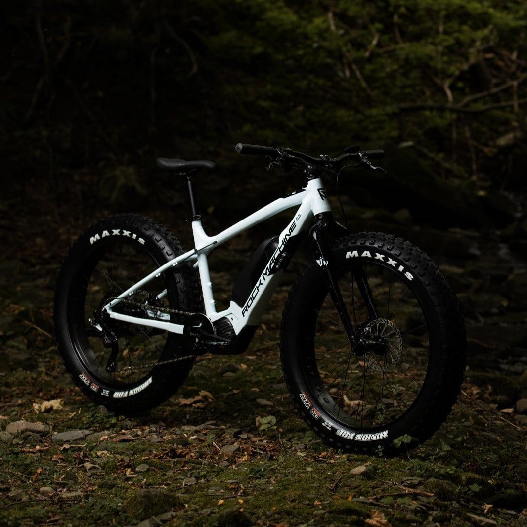 SĂHKĂPYĂRĂT.NET Rock Machine Pyöry E50R e-fatbike, jossa on paksut Maxxis-renkaat ja Shimano EP6 -moottori, seisoo kivikkoisella, lehtien peittĂ€mĂ€llĂ€ metsĂ€npohjalla, jota kehystÀÀ tumma, sumea puiden tausta.