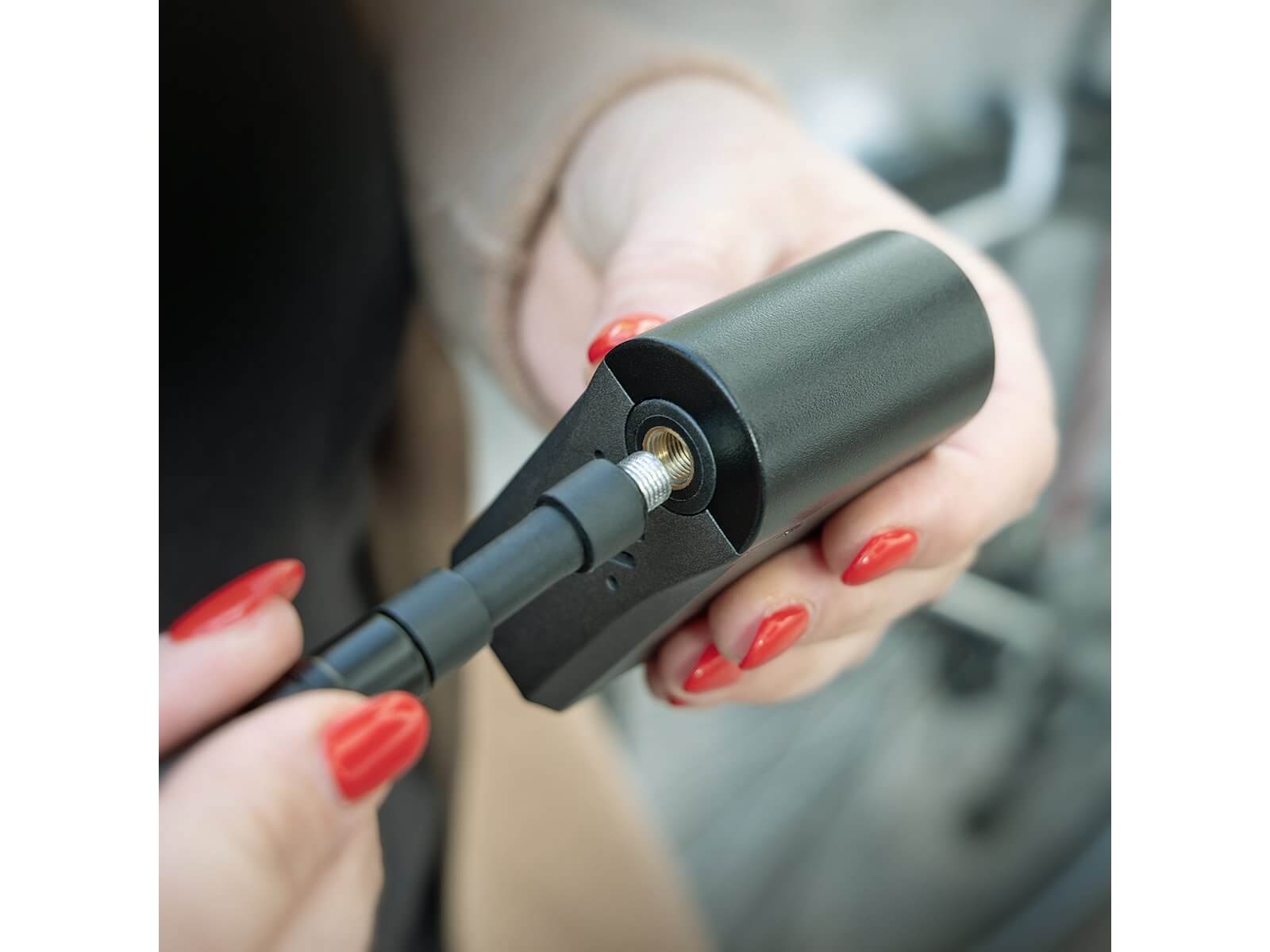 Henkilö, jolla on punaiseksi maalatut kynnet, liittää mustan kaapelin SKS elektroniseen pumppuun 8,3 bar / 120 psi, pitäen laitetta toisessa kädessä ja kaapelia toisessa kädessä.