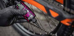 Mustaan hansikkaaseen pukeutunut henkilö levittää Muc Off All weather voiteluainepullosta Muc off -pulloa oranssin polkupyörän ketjuun keskittyen ketjuun ja Muc Off All weather voiteluainepulloon.