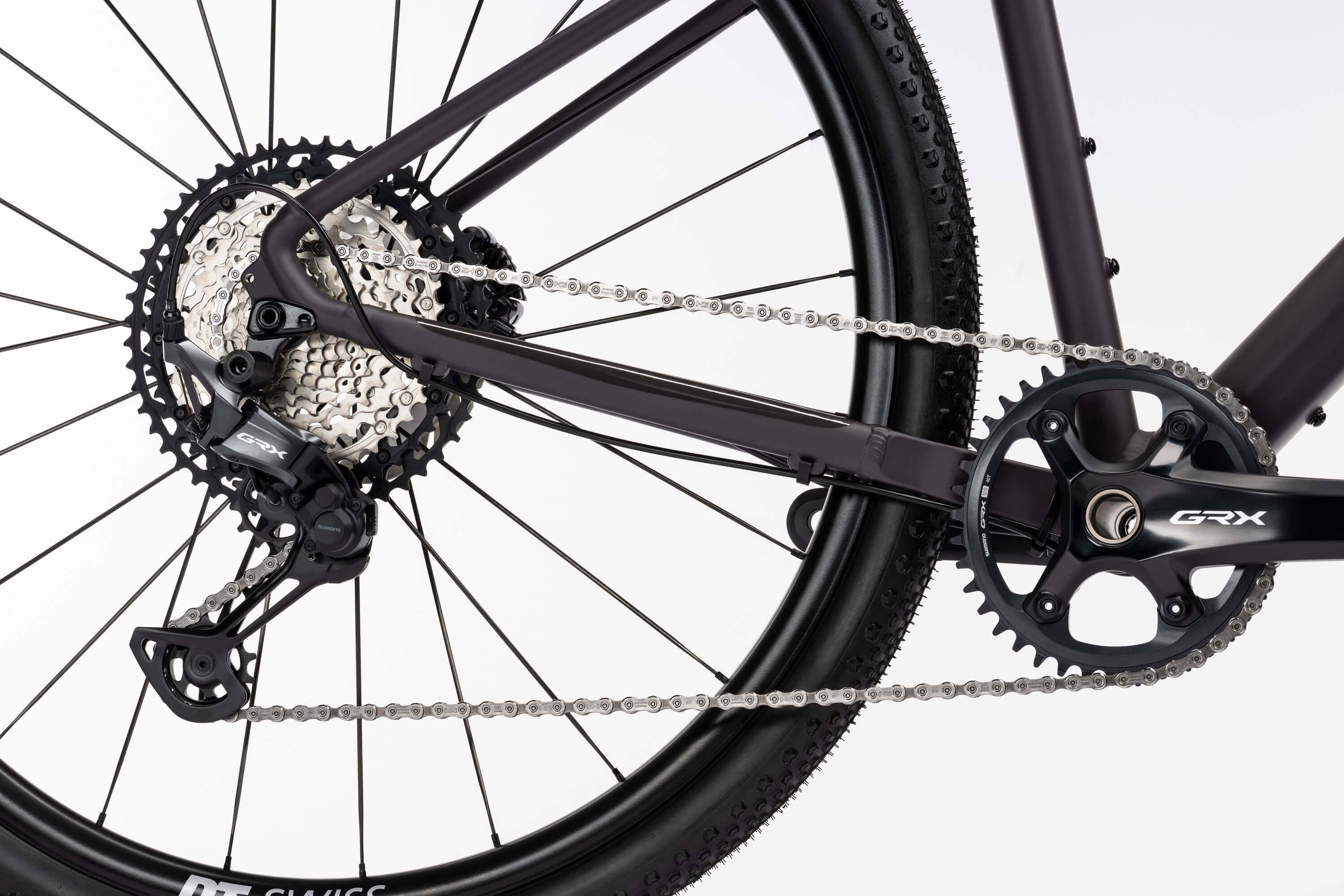 Lähikuva Ghost Asket Advancedin takapyörästä, jossa on Shimano GRX -kasetti, ketju, vaihteisto ja kampiakseli valkoista taustaa vasten. Korostaa Ghost-pyörän voimansiirtoa ja osaa rungosta.