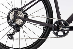 Lähikuva Ghost Asket Advancedin takapyörästä, jossa on Shimano GRX -kasetti, ketju, vaihteisto ja kampiakseli valkoista taustaa vasten. Korostaa Ghost-pyörän voimansiirtoa ja osaa rungosta.