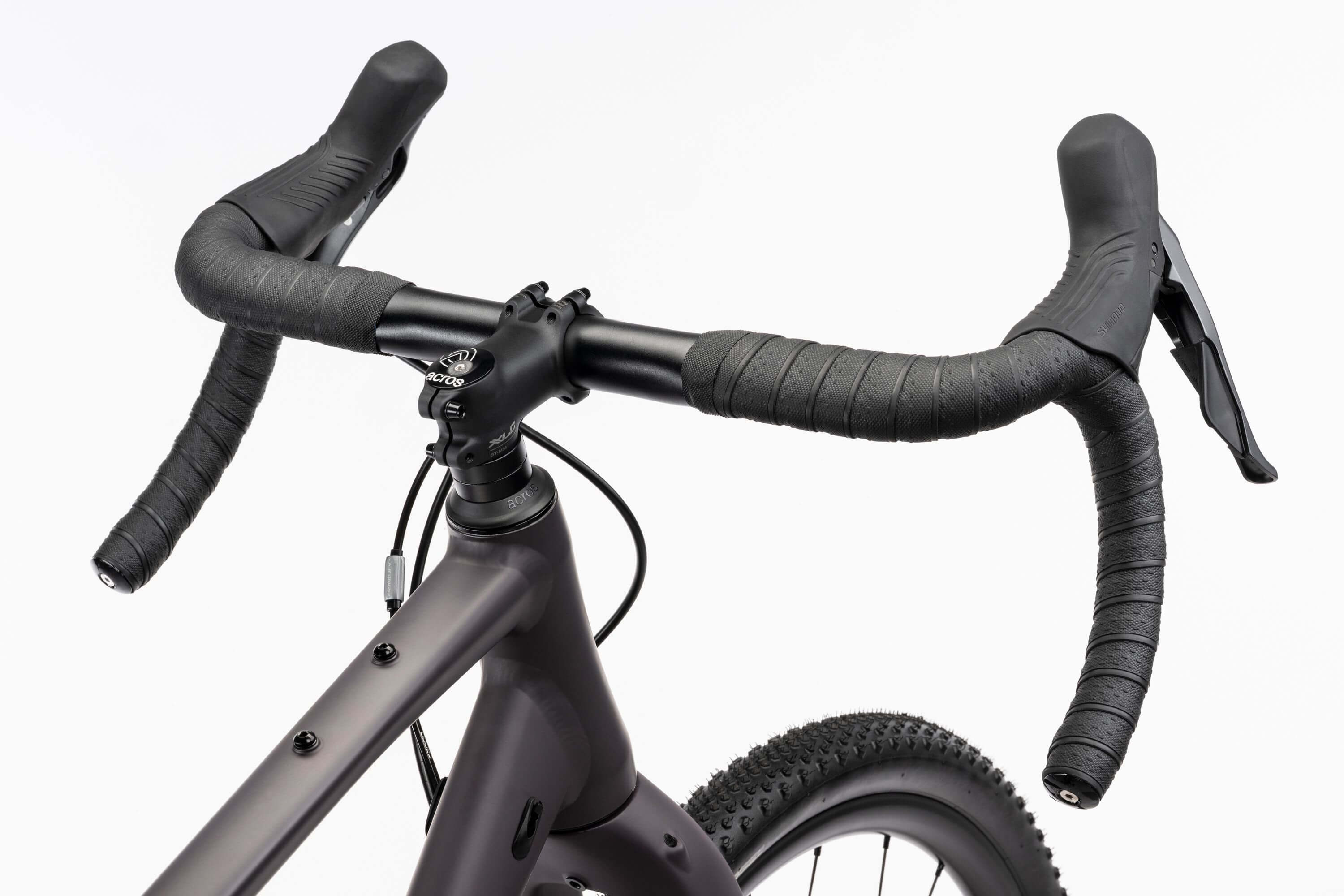 Lähikuva Ghost Asket Advanced -sorapyörän mustasta pudotetusta ohjaustangosta, jossa on käärityt kahvat ja joka on asennettu mattaharmaaseen Ghost-runkoon. Shimano GRX -jarruvivut ja nupukkarenkaat näkyvät valkoista taustaa vasten.