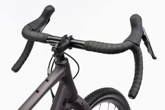 Lähikuva Ghost Asket Advanced -sorapyörän mustasta pudotetusta ohjaustangosta, jossa on käärityt kahvat ja joka on asennettu mattaharmaaseen Ghost-runkoon. Shimano GRX -jarruvivut ja nupukkarenkaat näkyvät valkoista taustaa vasten.