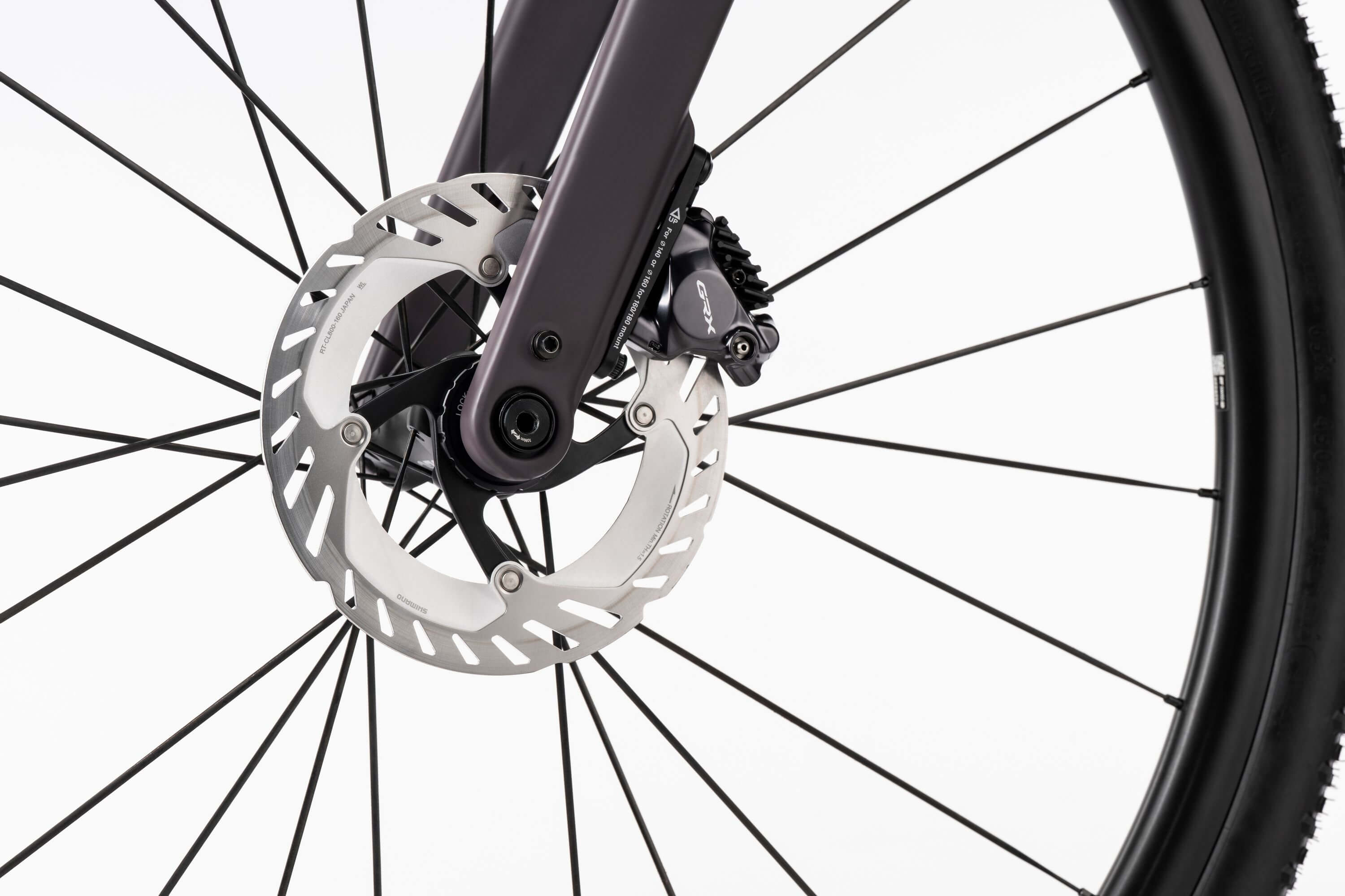 Lähikuva Ghostin Ghost Asket Advanced -etupyörästä, jossa näkyy hopeinen Shimano GRX -levyjarrulevy, musta jarrusatula ja mustat pyöränkehät valkoista taustaa vasten.