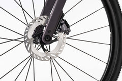 Lähikuva Ghostin Ghost Asket Advanced -etupyörästä, jossa näkyy hopeinen Shimano GRX -levyjarrulevy, musta jarrusatula ja mustat pyöränkehät valkoista taustaa vasten.