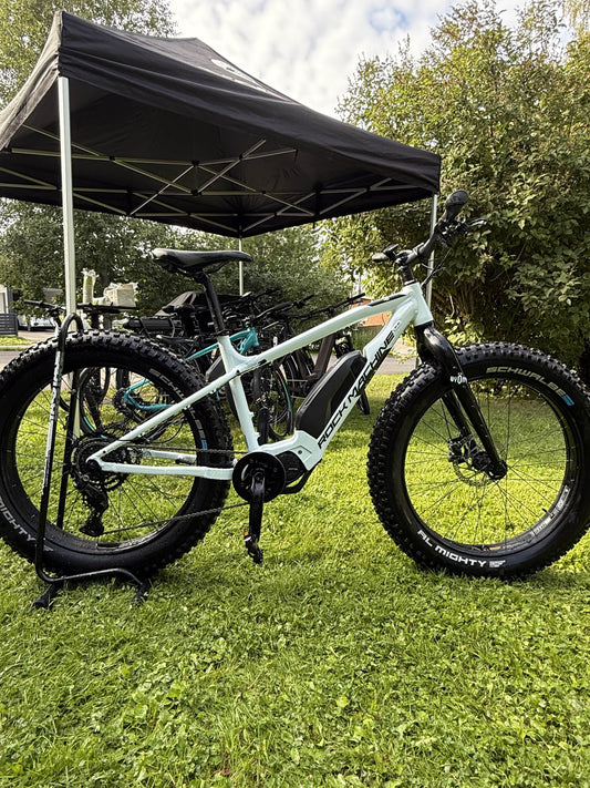 Valkoinen Rock Machine E50R esittelypyörä (vain 43km ajettu) SÄHKÖPYÖRÄT.NET:stä läskirenkailla näkyy nurmikolla mustan katoksen alla, muiden pyörien ja vihreiden lehtien ympäröimänä.