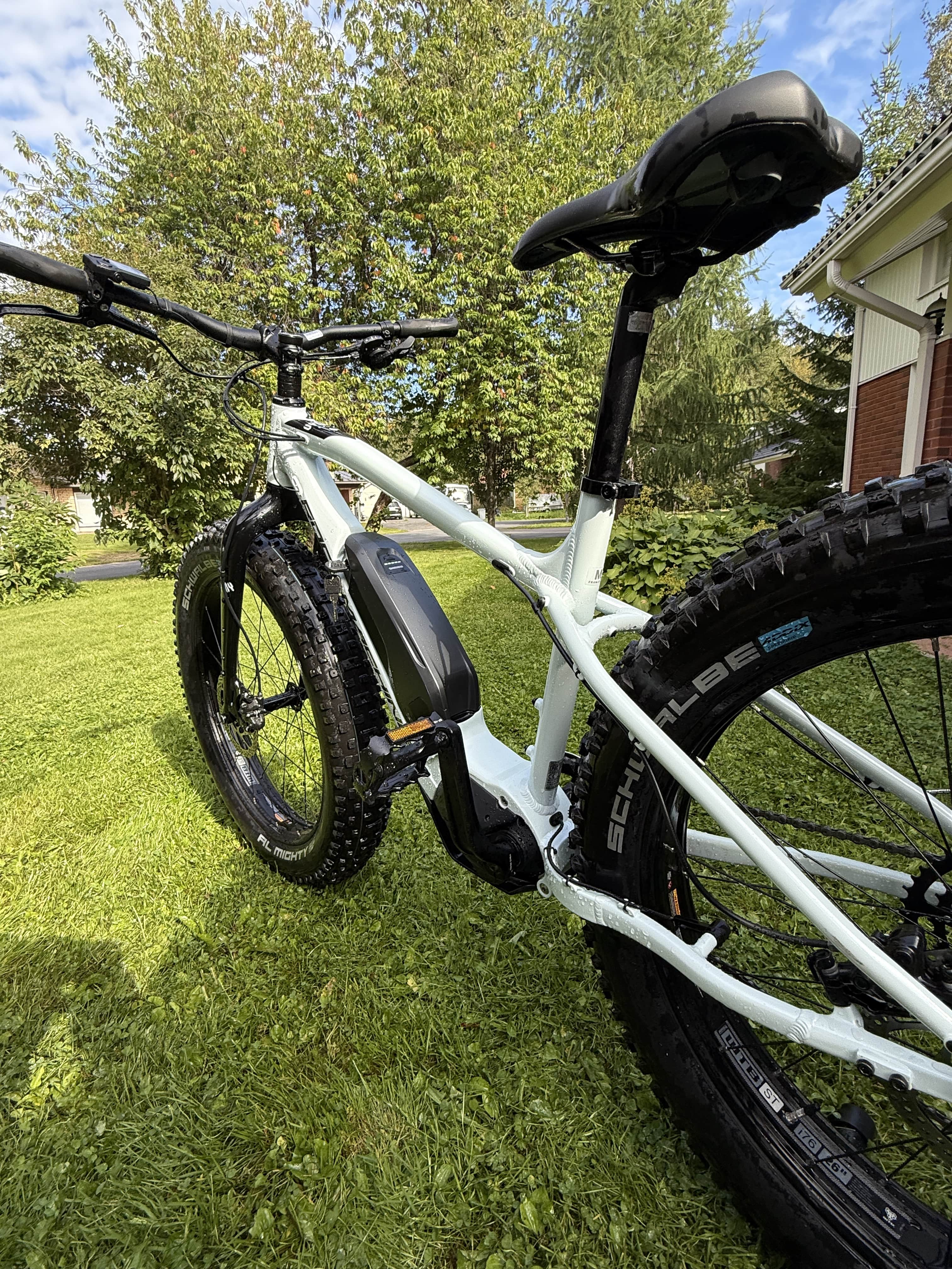 SÄHKÖPYÖRÄT.NET-sivuston matalan kuvakulman lähikuva Rock Machine E50R esittelypyörästä (vain 43 km ajettu), jossa on paksut renkaat ja joka on pysäköity nurmikolle; taustalla näkyy puita ja osa talosta osittain pilvisen taivaan alla.