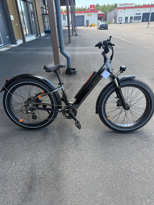 Musta SÄHKÖPYÖRÄT.NET Rad Power Bikes Radrhino 6 Plus Step Thru (käytetty, vain 48km!) leveillä renkailla on pysäköity asfaltille modernin lasirakennuksen lähelle. Taustalla on lähes tyhjä parkkipaikka ja teollisuusrakennuksia.