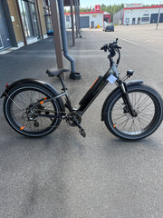 Musta SÄHKÖPYÖRÄT.NET Rad Power Bikes Radrhino 6 Plus Step Thru (käytetty, vain 48km!) leveillä renkailla on pysäköity asfaltille modernin lasirakennuksen lähelle. Taustalla on lähes tyhjä parkkipaikka ja teollisuusrakennuksia.