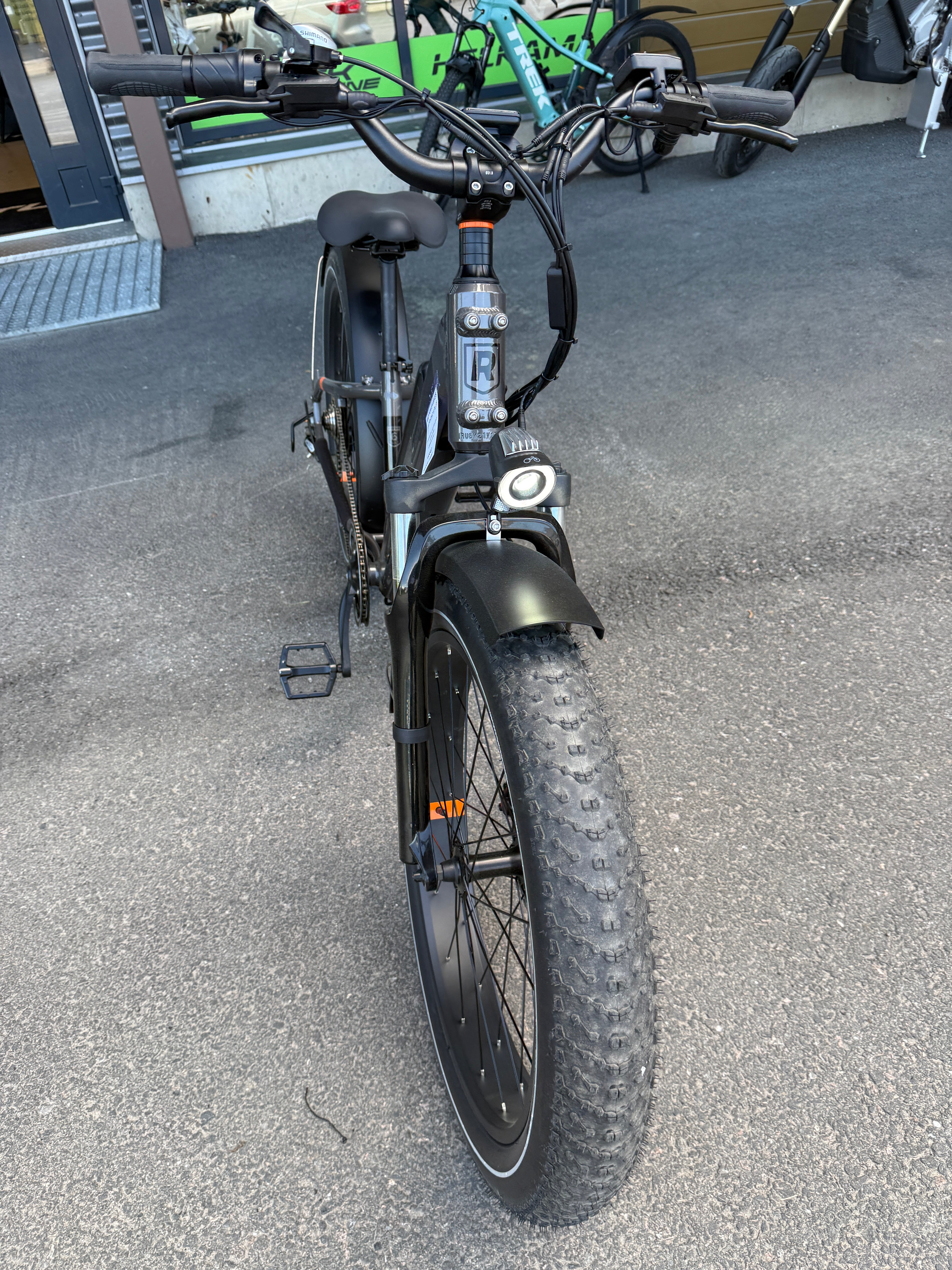 Musta SÄHKÖPYÖRÄT.NET Rad Power Bikes Radrhino 6 Plus Step Thru sähköpyörä (käytetty, vain 48km!), jossa on paksut renkaat, etuvalo ja pullonpidike, on pysäköity asfaltille rakennuksen lähelle; sen takana näkyy muita pyöriä.