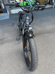 Musta SÄHKÖPYÖRÄT.NET Rad Power Bikes Radrhino 6 Plus Step Thru sähköpyörä (käytetty, vain 48km!), jossa on paksut renkaat, etuvalo ja pullonpidike, on pysäköity asfaltille rakennuksen lähelle; sen takana näkyy muita pyöriä.