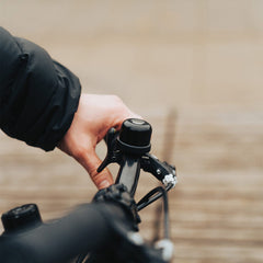 Lähikuva kädestä polkupyörän ohjaustangossa näyttää pyöräkellon ja jarruvivun. Pyöräilijällä on tumma takki. Sumeassa taustalla SÄHKÖPYÖRÄT.NET Pedalfinder pyörän GPS-paikannin mahdollistaa pyörän seurannan.
