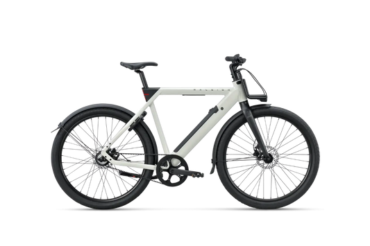 RALEIGH ONE hihnavetoinen sähköpyörä Raleighilta on moderni valkoinen kaupunkisähköpyörä, jossa on mustat pyörät, suojukset ja ohjaustanko. Valkoisella taustalla sivulta kuvattuna siinä on tukeva runko ja integroitu akku.