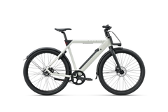 RALEIGH ONE hihnavetoinen sähköpyörä Raleighilta on moderni valkoinen kaupunkisähköpyörä, jossa on mustat pyörät, suojukset ja ohjaustanko. Valkoisella taustalla sivulta kuvattuna siinä on tukeva runko ja integroitu akku.