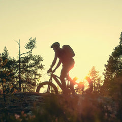 Kypärä ja reppu päässään olevan henkilön siluetti ajaa Tunturi EMAX *SUOMEN EDULLISIN* SÄHKÖPYÖRÄT.NETin Tunturi EMAX -pyörällä auringonlaskun polulla, taustalla puita ja auringonvalo paistaa pyörien läpi.