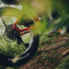 Lähikuva Tunturi EMAX *SUOMEN EDULLISIN* fatbiken takapyörästä ja vaihteista metsäpolulla, harmaasukkainen ajaja polkee; auringonvalo suodattuu vehreyden läpi. Saatavana SÄHKÖPYÖRÄT.NETistä.